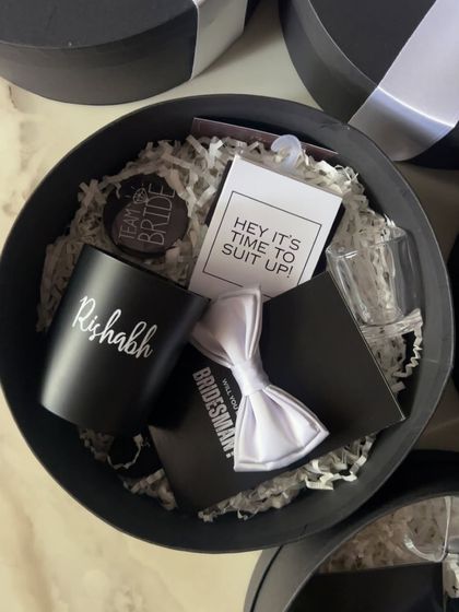 An overhead view of a round groomsmen proposal box. It contains a personalized mug, a bow tie card, a shot glass, and a "Team Bride" badge for the bridesman.