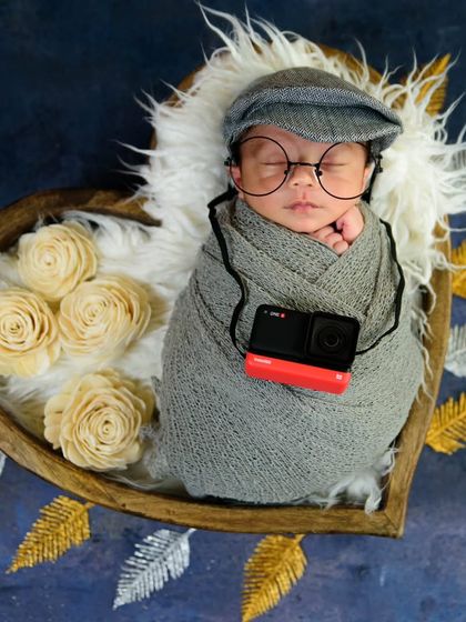 A future photographer in the making! This fun setup includes a newborn with a flat cap, glasses, and a miniature camera, nestled in a heart-shaped bowl.