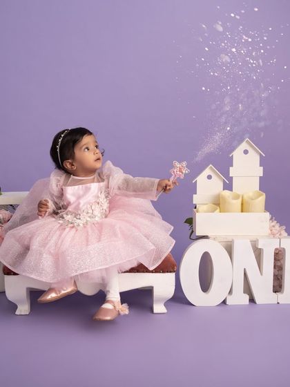 The magic of turning one. This little girl, dressed as a fairy, waves her wand in a dreamy purple-themed first birthday setup, complete with glitter and flowers.