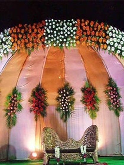A semi-circular stage backdrop decorated with panels of orange and white flowers. The draped fabric and floral bouquets create a classic look for a reception or sangeet night.
