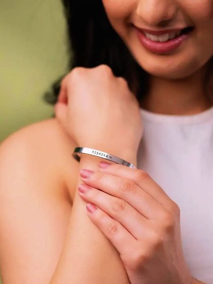 A smiling woman adjusts her silver cuff bracelet, engraved with the name 'RISHIKAM'. The focus is on the personal connection and happiness the jewelry brings.