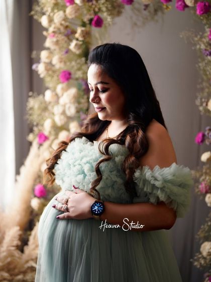 A gentle and serene portrait. The mother-to-be looks down at her bump with love, framed by a beautiful floral arch in the studio.