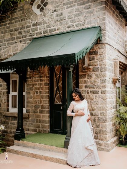 A beautiful portrait of the bride in her white engagement lehenga, posing in front of a stone building.