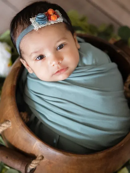 A close-up of a newborn's beautiful face, wrapped in a soft teal swaddle. The delicate headband adds a touch of detail to this simple and sweet portrait in a wooden bowl.