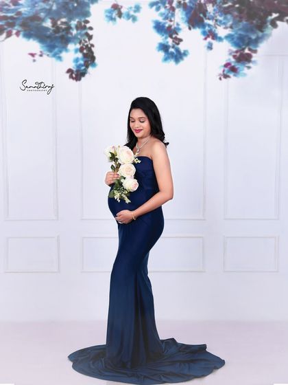 A solo portrait of a mother-to-be in a deep blue gown, holding a bouquet of white roses. The simple background and floral accents create a classic and elegant image.