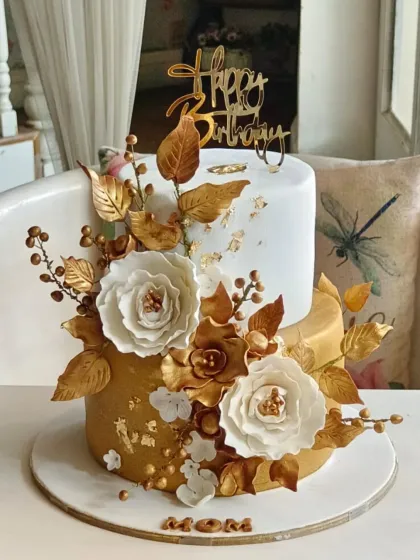 An opulent two-tier cake in white and gold, lavishly decorated with handcrafted white and gold sugar flowers and foliage.