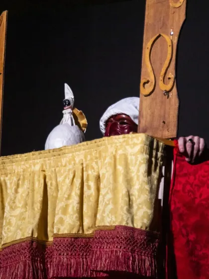 A close-up of a masked character peeking from behind the curtain in 'Io, Pulcinella', creating a moment of suspense and comedy.