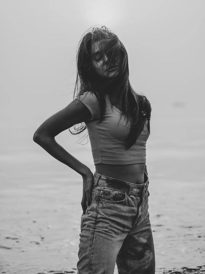 A full-body black and white shot on the beach, with the model's hair obscuring her face. It creates a sense of mystery and mood.