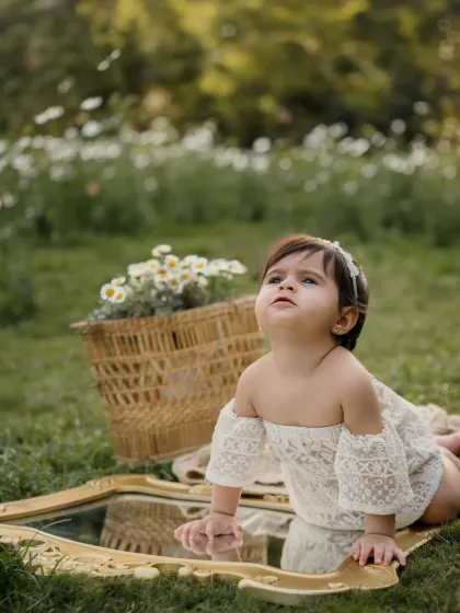 A baby girl in a white lace romper crawls over a mirror in a field, surrounded by flowers. A unique and artistic outdoor portrait.
