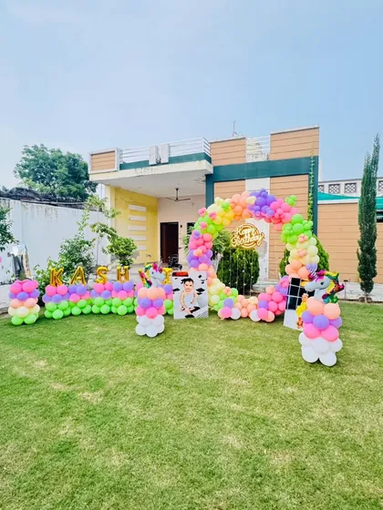 A magical unicorn-themed birthday party in an outdoor garden setting. The setup includes a rainbow balloon arch, unicorn cutouts, and foil balloons spelling out the birthday girl's name, "Kashvi."