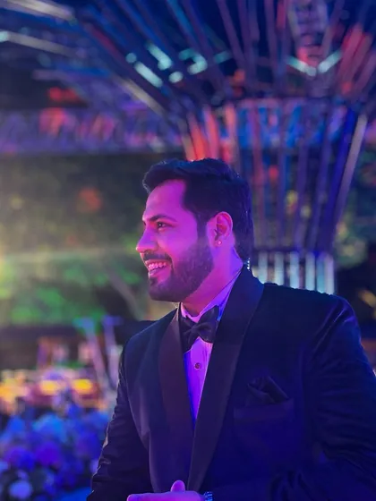 A profile shot, smiling, with the vibrant lights of the Sangeet in the background. Capturing the mood of the evening.