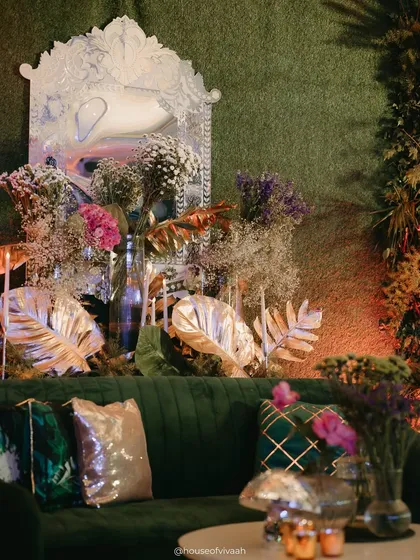 A luxurious lounge corner with an emerald green velvet sofa, metallic cushions, and a distorted mirror, set against a backdrop of gilded leaves and florals for a touch of surreal glamour.