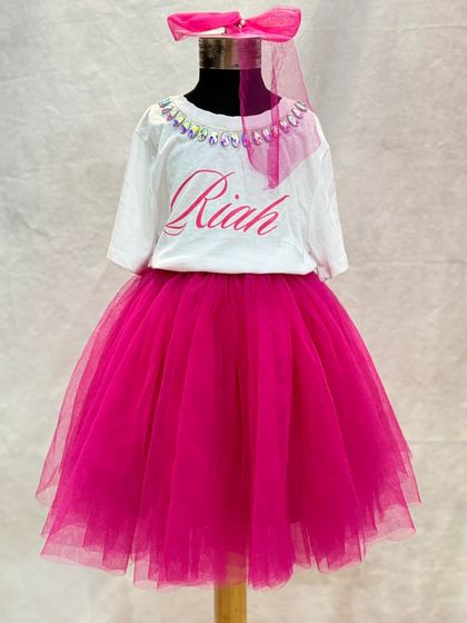 This custom birthday outfit is all about personal touches. The t-shirt features her name, "Riah," and a sparkling neckline, perfectly complementing the vibrant pink tulle skirt and matching hair accessory.