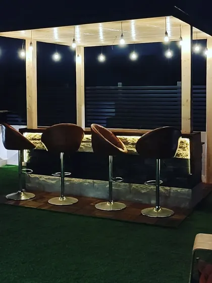 A beautiful rooftop bar at night. The bar is set under a pinewood-clad pergola and illuminated by hanging Edison bulbs for a warm, inviting glow.