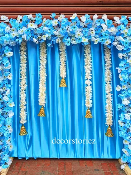 A serene blue and white themed ready-to-install backdrop for Ganpati. The design features a blue satin curtain, hanging jasmine garlands with temple bells, and a frame of artificial blue and white flowers.
