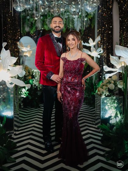 A full-length portrait of a couple ready for their Sangeet night. The striking black-and-white patterned floor and floral decor create a modern and chic backdrop for their glamorous outfits.