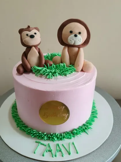 A sweet pink cake with hand-molded fondant animals, a lion and a monkey, sitting on a patch of green grass. Perfect for a little one's birthday.
