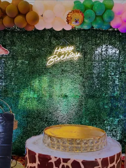 A fun and adventurous jungle-themed birthday party setup. We used a lush green foliage wall as a backdrop, accented with a neon 'Happy Birthday' sign, animal cutouts like a large giraffe, and themed cake stands.