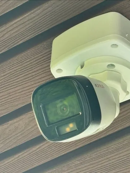 This CP Plus bullet camera is mounted under an eave on a textured exterior wall in GK-1. This placement protects the camera from direct rain and provides a clear view of the area below.