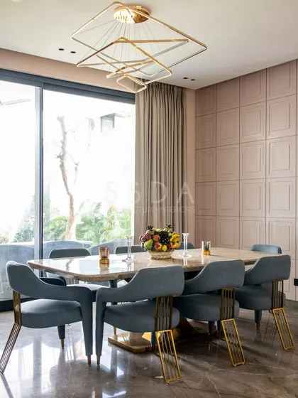 The dining room at 'House Retold' features a marble-topped table and sculptural chairs with gold accents. A feature wall with geometric panelling adds texture, while large glass doors connect the space to the garden.