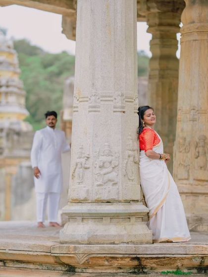 A portrait focusing on the woman in her beautiful white and red saree, leaning against a carved stone pillar. Her partner stands softly in the background, creating a sense of depth.