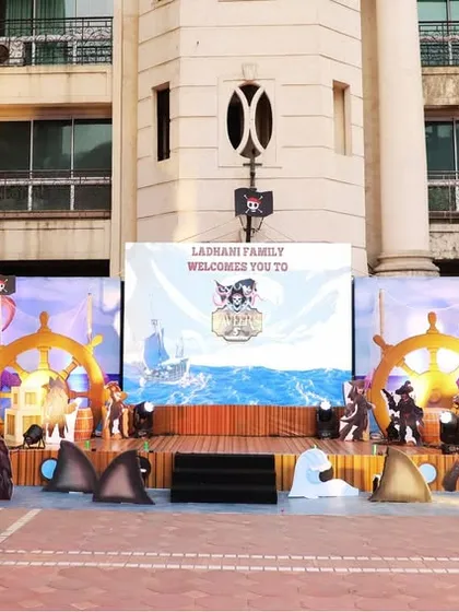 A full stage setup for a "Pirates and Sea Monsters" themed birthday party. The backdrop features a seascape, flanked by large, illuminated ship and sea monster props.