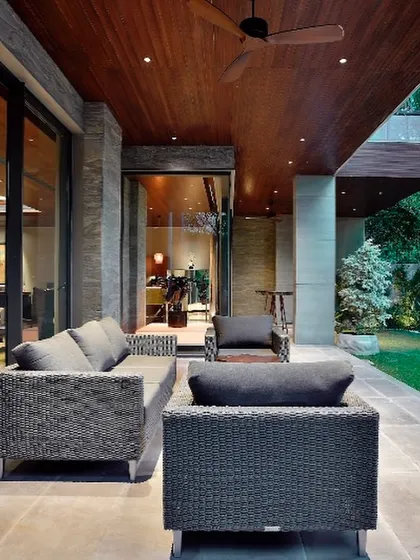 An outdoor seating area at the T House, tucked under a cantilevered upper floor. The space is defined by a warm wooden ceiling and stone flooring, creating a cozy, sheltered patio that opens directly onto the lawn.