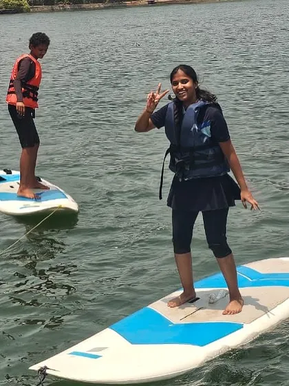 Two girls enjoy paddleboarding together, smiling and waving from their boards at the Karwar sailing camp.