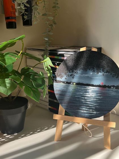 The round nightscape painting on an easel, next to a plant.