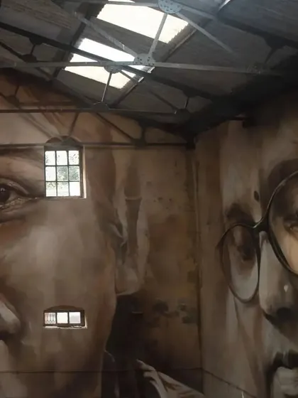 An interior view of the Sassoon Dock warehouse, featuring the monumental portraits of the local community by artist Guido Van Helten. The natural light from the skylights illuminates the faces.