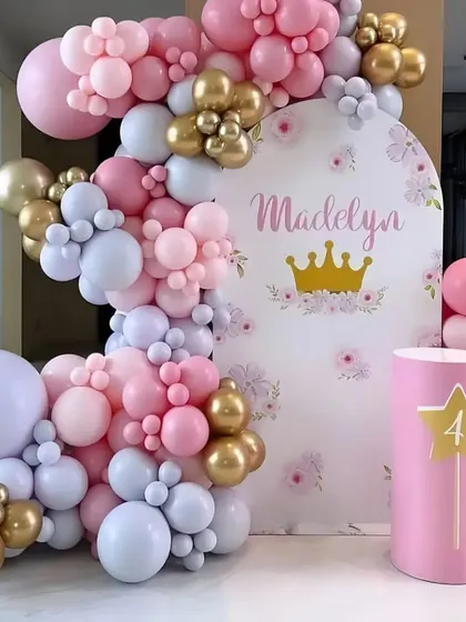 A princess-themed 4th birthday party. This decoration features a custom backdrop with a crown, a pink and purple balloon arch, and a pedestal for the cake.
