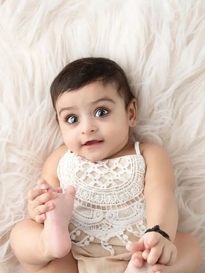 A baby girl in a lace outfit lies on a white fur rug, holding her foot.