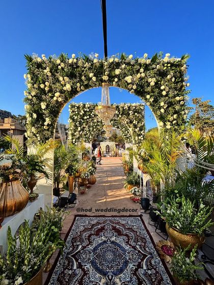 A breathtaking outdoor entrance with arches covered in white roses, leading down a path lined with greenery and traditional decor elements.
