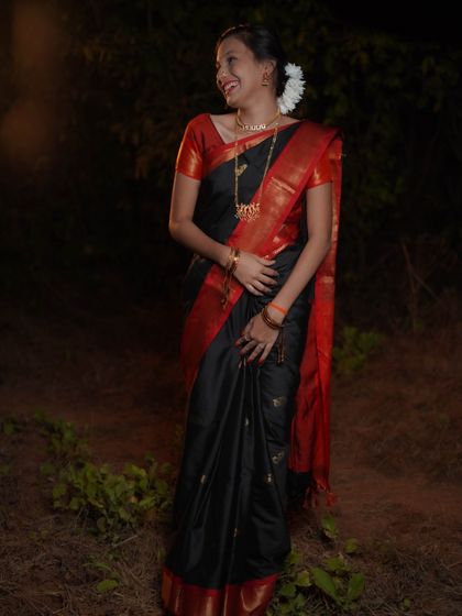 A candid, full-length shot of a woman in a traditional black and red saree for a Makar Sankrant celebration. Her natural laugh and the outdoor, rustic setting create a beautiful, unposed moment.
