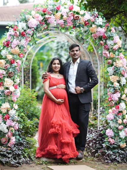 A beautiful couple's maternity portrait under a floral arch. The vibrant red of the ruffled gown stands out, while the partner's classic suit creates a timeless look.