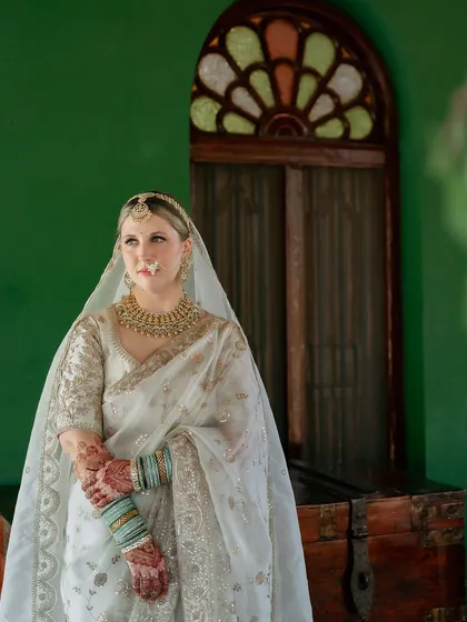 A bride in a white and gold saree, her face framed by her veil and a stained-glass window, a truly elegant and timeless portrait.