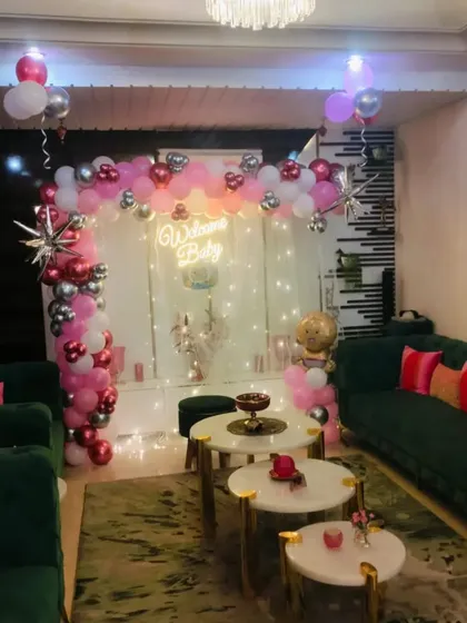 A sophisticated welcome baby setup in a living room, using a balloon arch in pink, white, and chrome red. The backdrop is accented with fairy lights and a "Welcome Baby" neon sign for a warm, glowing effect.