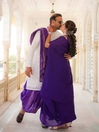 A romantic kiss shared in a sunlit palace corridor. The bride's deep purple saree and the groom's traditional white and purple attire create a striking and passionate image.