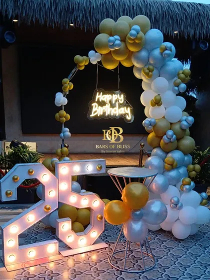 An outdoor 25th birthday celebration with a yellow, silver, and white color scheme. The decor features a balloon arch, a 'Happy Birthday' neon sign, and large marquee numbers that stand out beautifully against the evening sky.