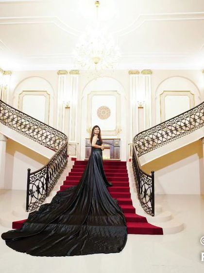 A breathtaking wide shot of a bride in a magnificent black gown with a long train, standing on the grand staircase of a palace-like studio set.