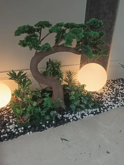 An elegant indoor garden created with a sculpted artificial bonsai tree, smaller plants, and a bed of black and white pebbles. The integrated globe lights provide a warm glow, turning this corner into a sophisticated feature.