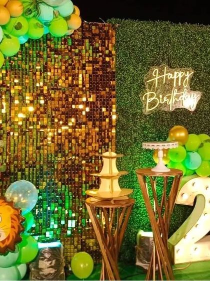 A close-up of our jungle theme decor, highlighting the mix of textures with a gold sequin panel next to a green foliage wall. The setup includes a neon 'Happy Birthday' sign and rustic wooden stands.