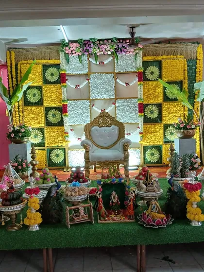 A beautiful baby shower setup with a checkerboard backdrop of yellow marigolds and white jasmine. The stage is set on a green lawn and includes a full display of offerings with peacock figures.