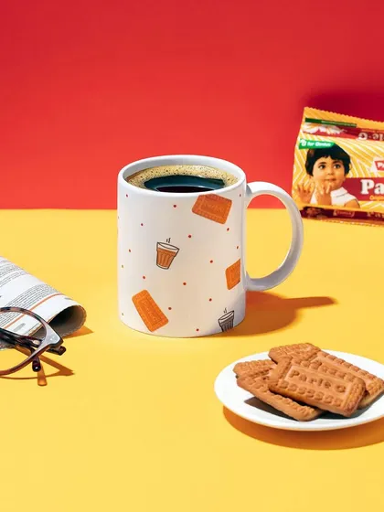 A nostalgic and classic setup for Parle-G's merchandise line. This image features their branded mug alongside the iconic biscuits, a newspaper, and glasses, evoking the feeling of a perfect morning tea time.