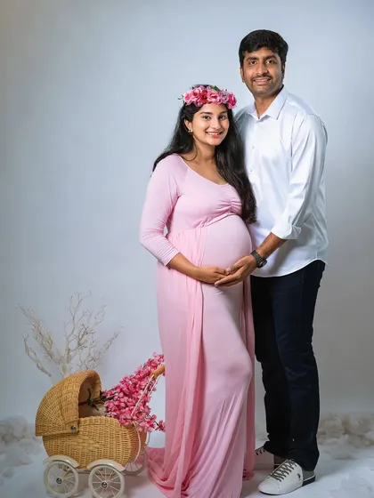 A sweet and classic studio portrait of an expecting couple. The mom-to-be wears a soft pink gown and floral crown, standing next to her partner with a vintage-style pram prop nearby.