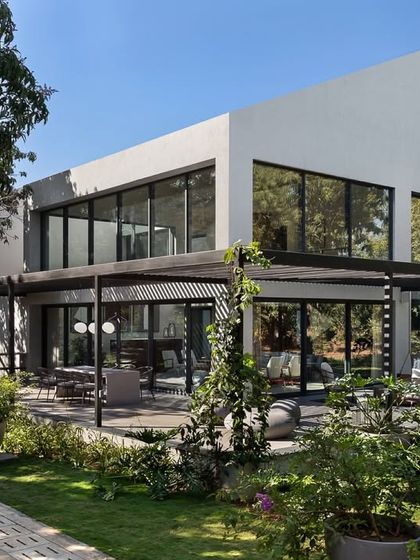 The exterior facade of the Mango Orchard House is a composition of solid white walls and large glass voids. A black metal pergola extends from the ground floor, creating a shaded outdoor living area that integrates with the garden.