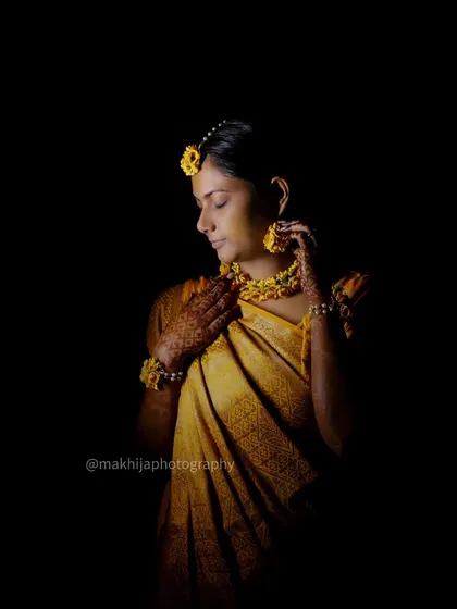 A dramatic portrait of the bride during her Haldi ceremony. The dark background makes the yellow of her saree and floral jewelry pop, creating a stunning and artistic image.