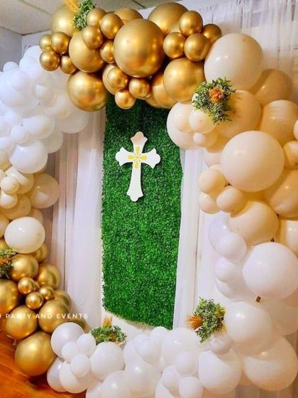 An elegant baptism decoration using a balloon ring against a faux grass panel. The white and gold balloons are accented with small touches of greenery.