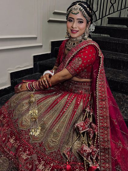 A full-length shot of a bride in a stunning red and gold lehenga. The makeup is classic and elegant, with a neat bridal bun to complete the look.