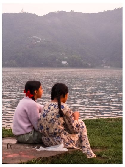 Two young girls with braided hair sit by a lake, looking out at the mountains in the distance. It's a peaceful, contemplative moment captured in soft light.